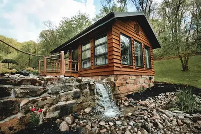 a log cabin with a waterfall in front of it at Lake with Dock and Hot Tub Hocking Hills Cabin in Rockbridge