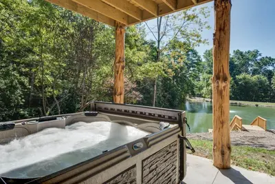 a hot tub under a wooden pergola next to a river at Stone Lake Lodge - Lakeside Cabin - Stocked Pond in Coshocton