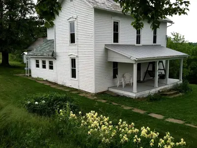 a white house with a porch and chairs on it at 1800s Farmhouse Cottage Rental on a 230 Acre Farm in Muskingum County, Ohio in Frazeysburg